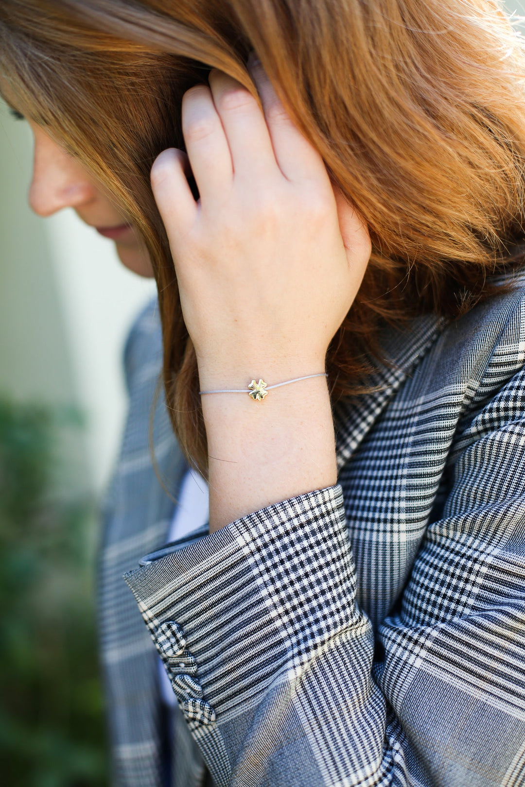 A person with brown hair, wearing a checkered blazer, touches their hair. On their wrist rests a delicate 14k gold Wish Birthday bracelet by LuckyFeather, adorned with a clover charm. The background is blurred with greenery visible, enhancing the serene moment.