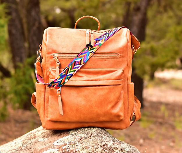 The Hailey Backpack - Brown by zzzLivFoxy rests on a rock, showcasing multi-functional pockets and a vibrant, patterned adjustable strap. The blurred greenery in the background hints at an outdoor setting.