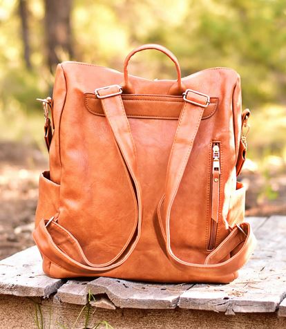 The Hailey Backpack by zzzLivFoxy, featuring a brown vegan leather finish with adjustable straps and multi-functional pockets, sits on a wooden surface outdoors. The blurred green foliage in the background suggests a natural setting.