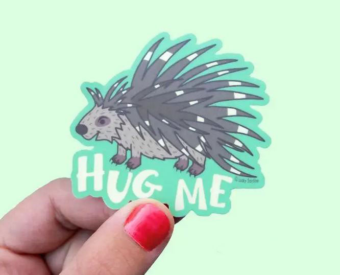 A hand with painted nails holds the adorable Hug Me Vinyl Sticker by Lucky Sardine, featuring a cute cartoon porcupine and the bold phrase "HUG ME." Set on a light green background, this charming accessory is designed to be both scratch-resistant and water-resistant for long-lasting durability.