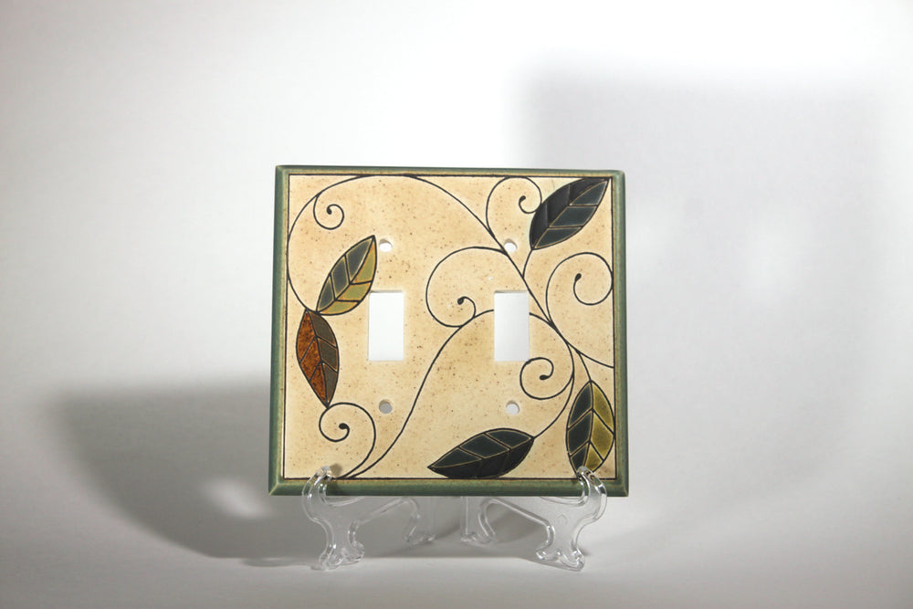 The "Double Mosaic Leaves" ceramic switch plate cover by ALL FIRED UP, crafted in Canada, showcases a stunning leaf and vine design. Elegantly presented on a clear stand against a simple backdrop, this rectangular cover includes two switch apertures, bringing a distinctive flair to any room.