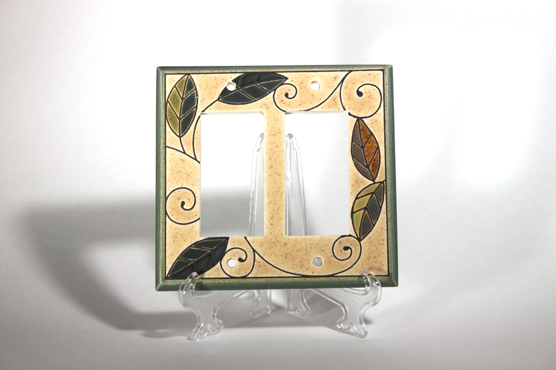 The Double Rocker Mosaic Leaves by ALL FIRED UP is a custom-made, decorative double light switch cover with a beige background, featuring green, brown, and black leaf patterns and swirls. Made in Canada, it is showcased standing upright on a clear display against a neutral backdrop.