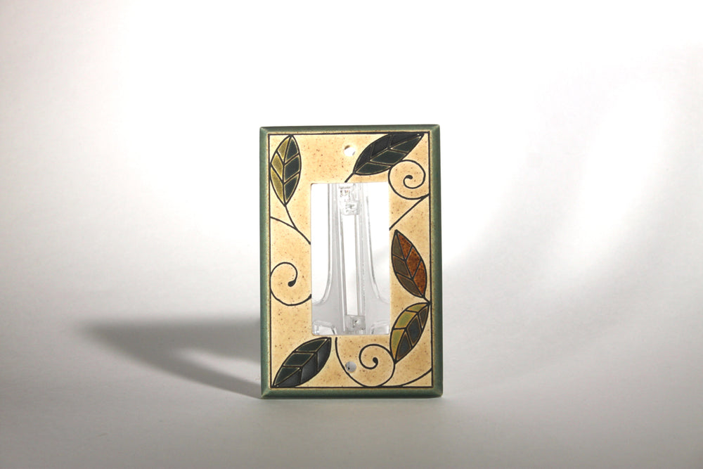 The Single Rocker Mosaic Leaves by ALL FIRED UP is a custom-made ceramic switch plate cover with a distinctive clear switch. It showcases an elegant design of black and gold leaves against a beige backdrop, crafted in Canada. The centered switch casts a subtle shadow on the white surface.