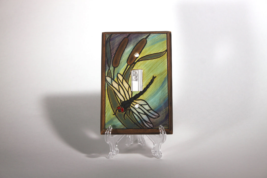 The Single Dragonfly Bulrush light switch cover, featuring a stained glass design of a dragonfly and cattails, is crafted by ALL FIRED UP in Canada and displayed on a clear plastic stand against a plain white background.