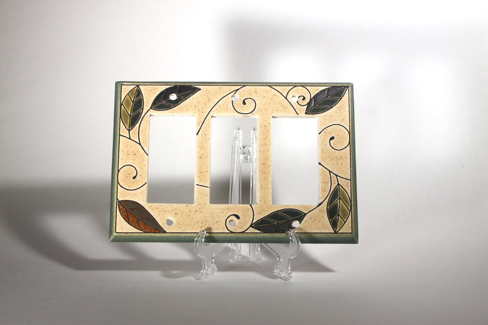 A Triple Rocker Mosaic ceramic switch plate cover from ALL FIRED UP stands on a clear plastic easel. Crafted in Canada, it features a beige background with green and brown leaves intertwined with vine-like black swirls. The decorative switch plate has openings for two rectangular and one circular switch.