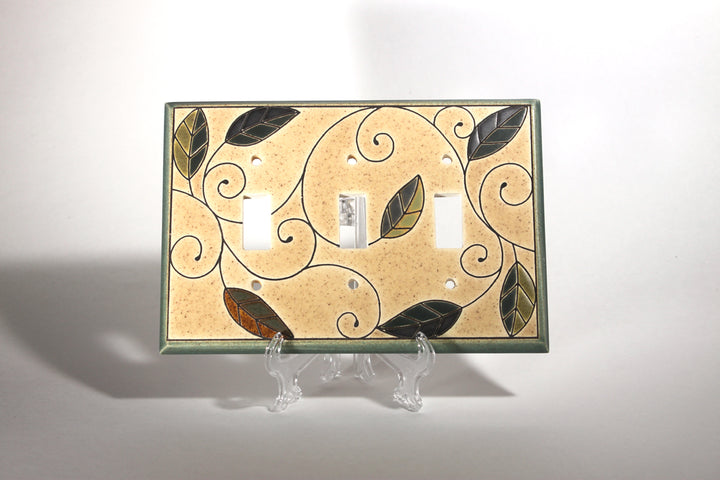 The "Triple Mosaic Leaves" switch plate cover by ALL FIRED UP is a custom-made ceramic piece featuring three openings. It showcases a beige background adorned with an ornate design of green and brown leaves connected by swirling vines. Crafted in Canada, the switch plate is displayed on a clear stand against a plain white background and comes with replacement screws included.