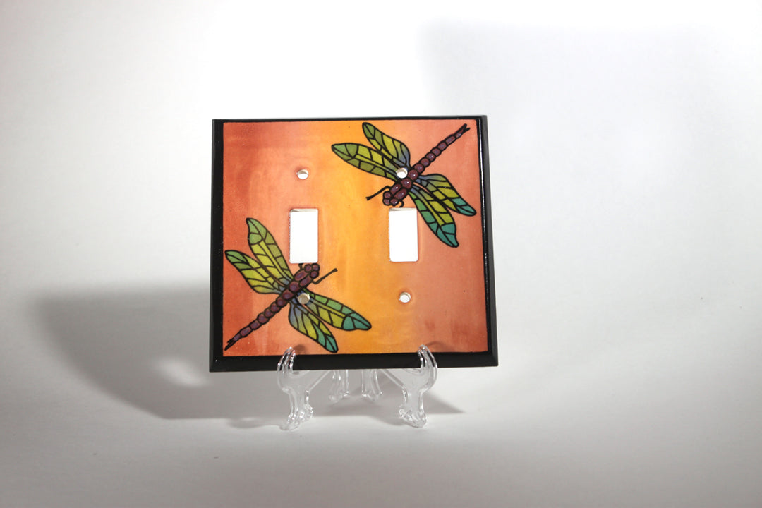 Displayed on a clear stand is the "Double Multicolor Dragonfly" switch plate cover by ALL FIRED UP. Crafted in Canada, this decorative ceramic piece features two dragonfly designs on an orange background, adding a vibrant touch to any room.