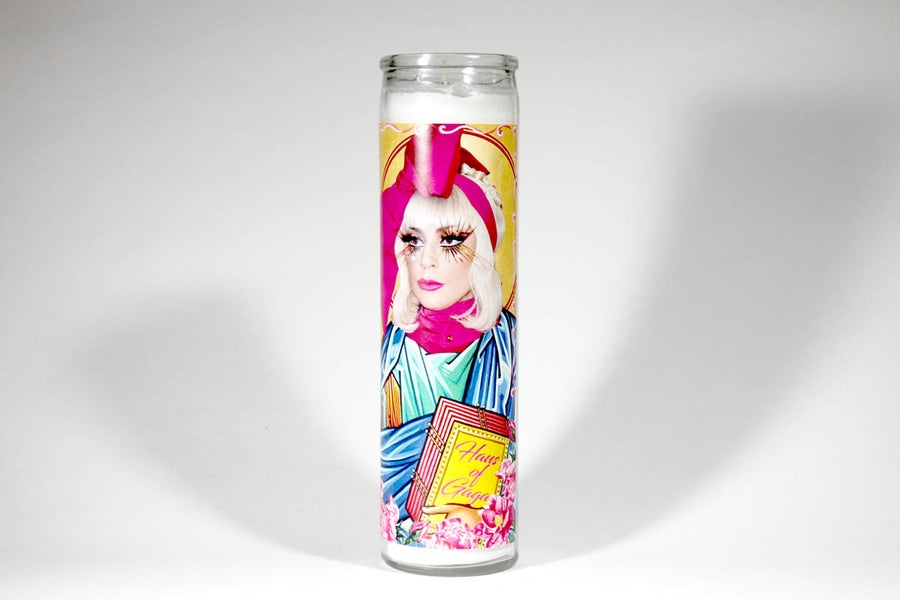 The Lady Gaga candle by BOBBYK BOUTIQUE is a white, unscented prayer candle showcasing Lady Gaga with a pink headdress, dramatic eye makeup, vibrant robes, and a book. Surrounded by floral elements and text, it makes for a unique gift.