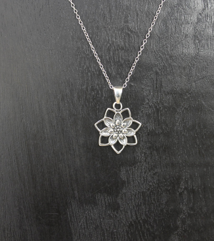 The Sterling Silver Lotus Bloom Pendant by Welman features a delicate chain and detailed floral design, making it perfect for everyday wear against a textured dark background.