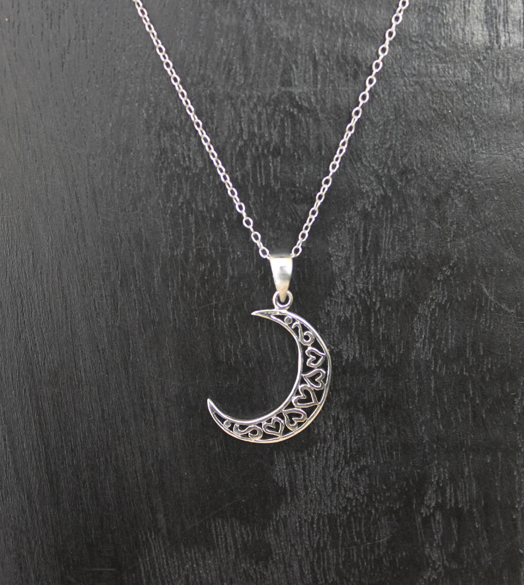 A delicate sterling silver necklace with a Heart Moon pendant by Welman is displayed against a dark background. The 925 Sterling Silver pendant features intricate filigree work, giving it an elegant and ornate appearance. The fine sterling silver chain is thin and complements the design of the pendant perfectly.