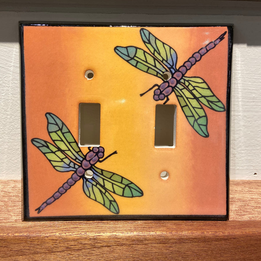 The Double Multicolor Dragonfly ceramic switch plate cover by ALL FIRED UP, crafted in Canada, features two dragonflies with green wings and purple bodies against a vibrant orange backdrop, ideal for adding a whimsical charm to any space.