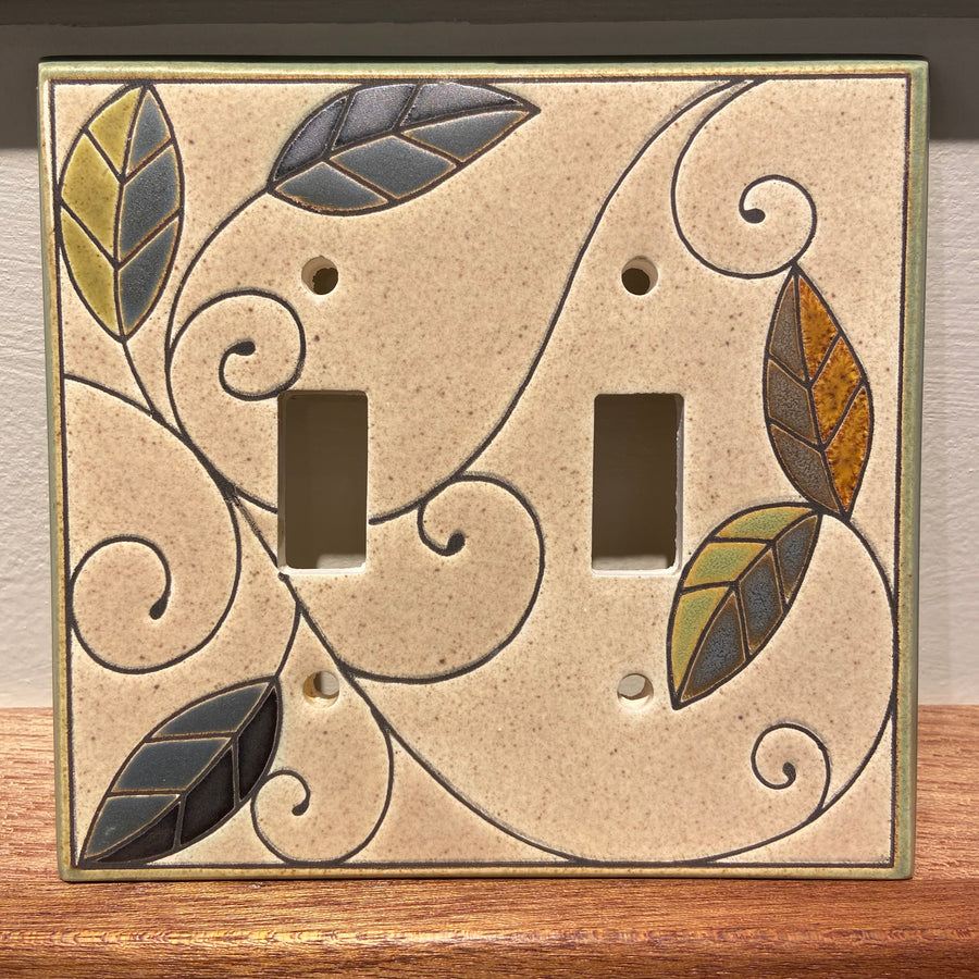 Introducing the "Double Mosaic Leaves" by ALL FIRED UP: a custom-crafted decorative light switch cover featuring dual openings. Adorned with vibrant leaves and swirling vine patterns, this ceramic plate is skillfully made in Canada and showcases shades of green, blue, and brown set against a beige backdrop.