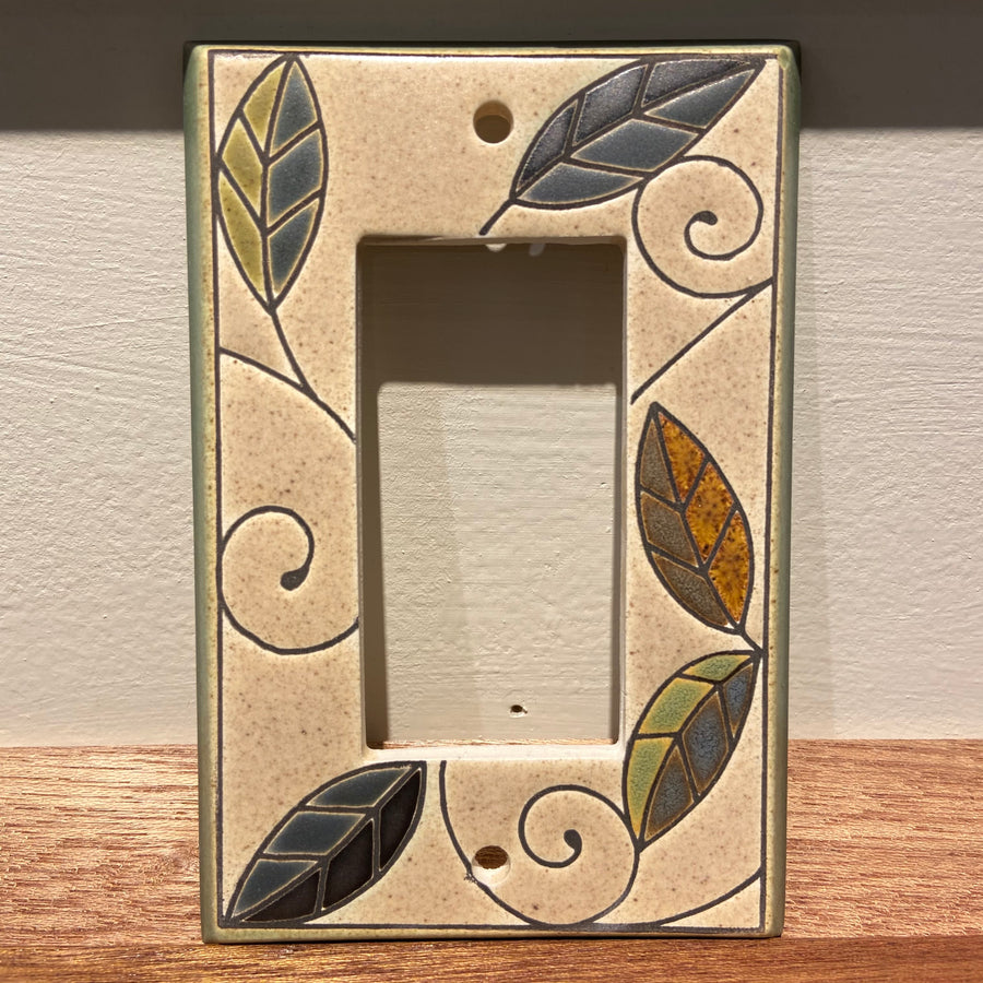 The Single Rocker Mosaic Leaves by ALL FIRED UP is a decorative ceramic switch plate cover with a rectangular cut-out. It features a beige background adorned with swirling lines and colorful leaf designs in green, blue, orange, and gray. This custom-made piece from Canada is placed on a wooden surface against a light wall.