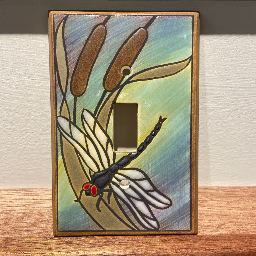 The "Single Dragonfly Bulrush" light switch cover by ALL FIRED UP features a charming decorative design with a dragonfly, complete with red eyes and transparent wings among cattails. Set against hues of blue, green, and brown, this Canadian-crafted wooden piece beautifully enhances any collection of ceramic switch plate covers.