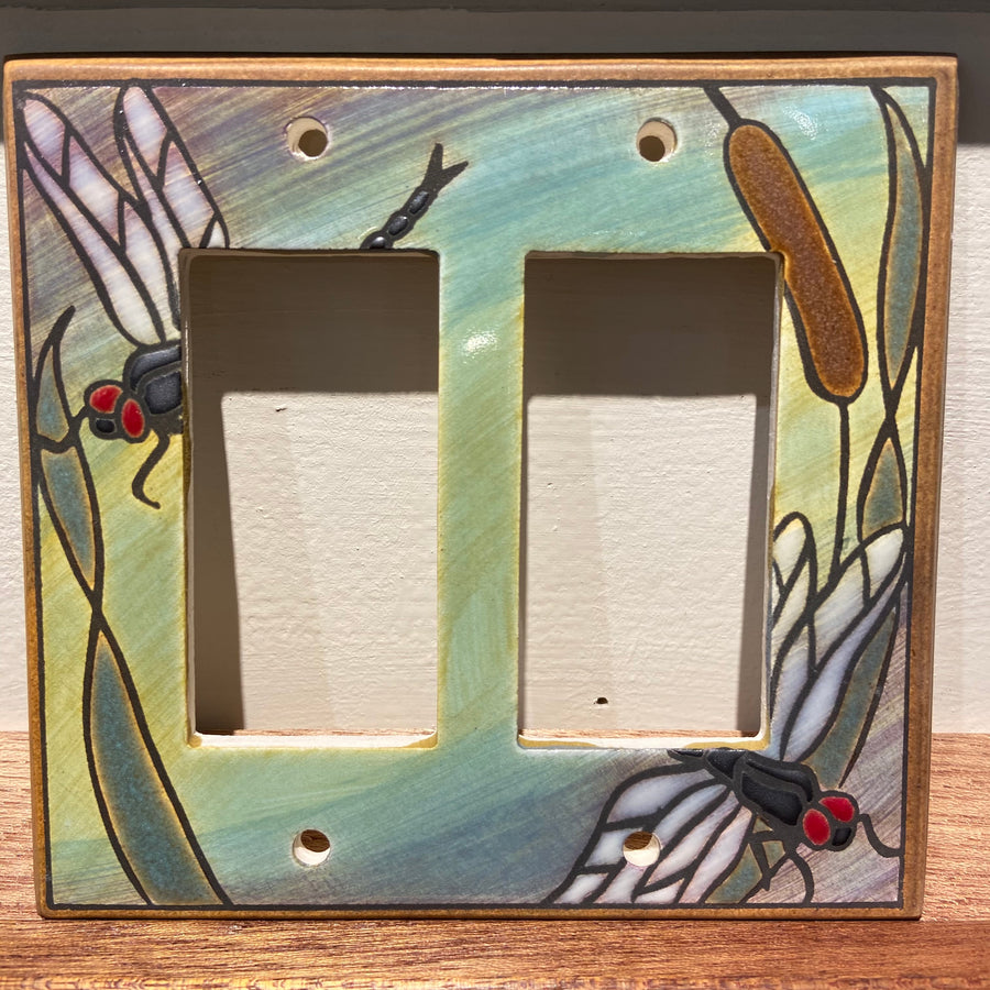 The Double Rocker Dragonfly Bulrush by ALL FIRED UP is a decorative ceramic switch plate cover made in Canada, featuring vibrant dragonflies and cattails on a blue-green background. It includes two rectangular openings for switches and comes with replacement screws for easy installation.