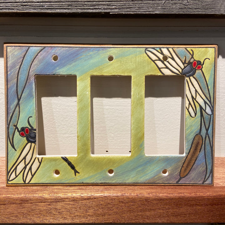 The Triple Rocker Dragonfly Bulrush switch plate cover from ALL FIRED UP is a decorative ceramic piece with hand-painted dragonflies on a green and blue background, featuring three rectangular switch openings. Crafted in Canada, it includes replacement screws for easy installation.