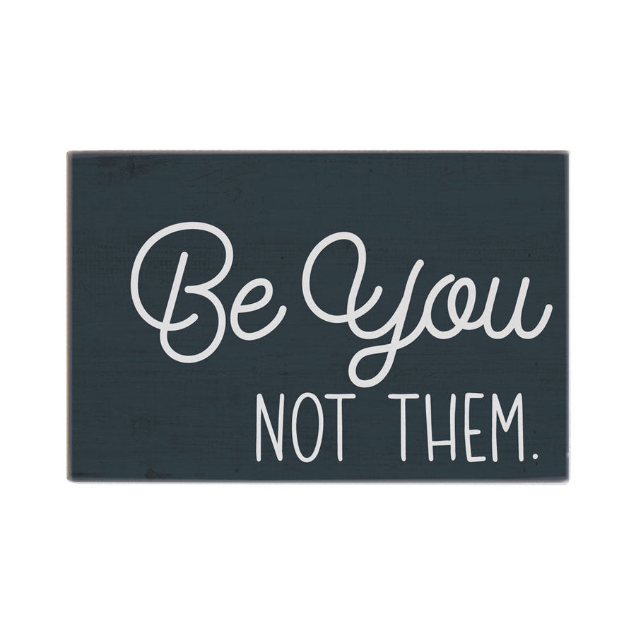 The "Be You Not Them" sign by SincereSurroundings is a dark green, handcrafted piece featuring white text that reads, "Be You NOT THEM." The solid background creates a striking contrast with the mix of cursive and block lettering. It's perfect for displaying on a shelf in Rock Rapids or as a standout piece in your home.