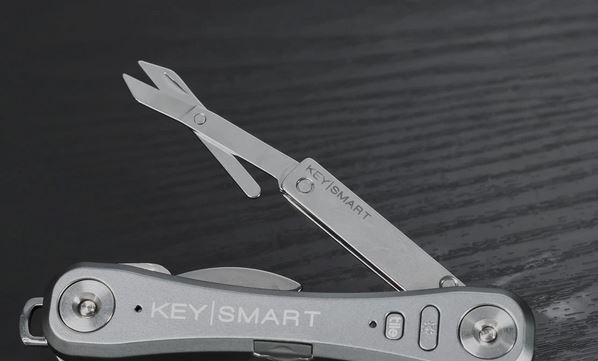 A compact multi-tool called "KeySmart NanoScissors" is displayed on a dark wooden surface. It features easy spring-action cut scissors extended and housed within a sleek, metallic casing, along with additional tools, including a small blade retracted within the durable stainless steel case.