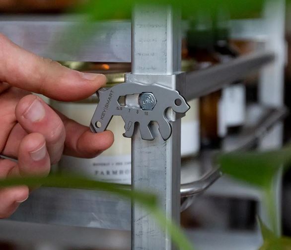 A hand holds a KeySmart Bear Keychain Multitool against a metal frame. This outdoor tool boasts various functions, including a ruler and hex wrenches. Green leaves partially obscure the view, highlighting its lightweight durability for your adventurous needs.