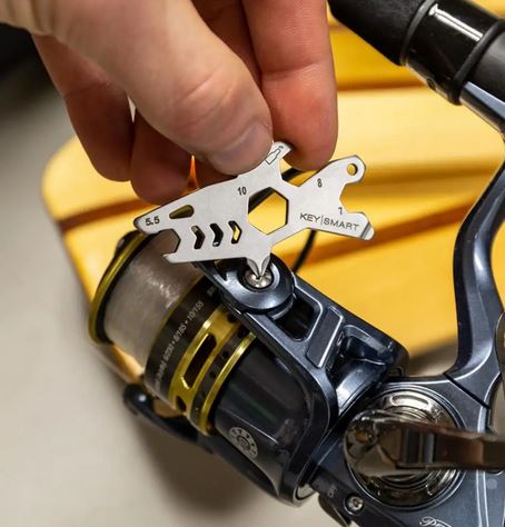 A hand is seen holding a lightweight, durable Shark Keychain Multitool from KeySmart next to a fishing reel. The multitool features several cutouts, including a wrench with measurements marked "5.5," "10," and "8." The fishing reel is mounted on a rod with a yellow and black spool.