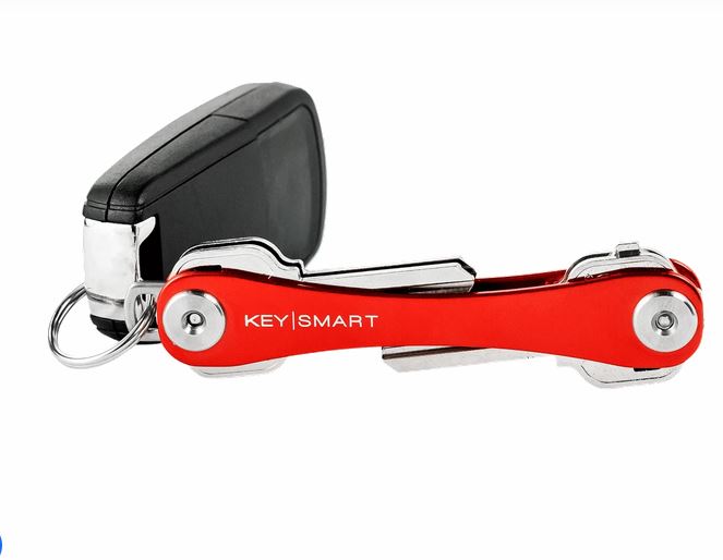 A red KeySmart Key Holder from the brand KeySmart is shown with a sleek design. It comfortably holds several organized keys and is attached to a black car key fob by a small metal ring. The word "KEYSMART" is printed on the red key holder in white lettering.