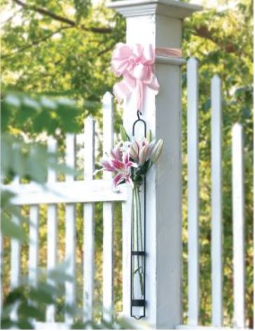 Featuring a versatile white wooden fence topped with an elegant pink ribbon bow, Couronne's Florence Tube Vase with Hanger Gift Box enhances the scene. Below, its unique design element—a metal holder crafted from recycled glass—cradles pink and white lilies against a lush backdrop of green foliage.