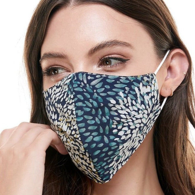 A person with long brown hair is wearing the zzzMiley and Molly Mask - Paint Stroke, which features a blue and white leaf pattern. This reversible face mask covers the nose and mouth, while elastic ear bands provide a snug fit against the plain white background.