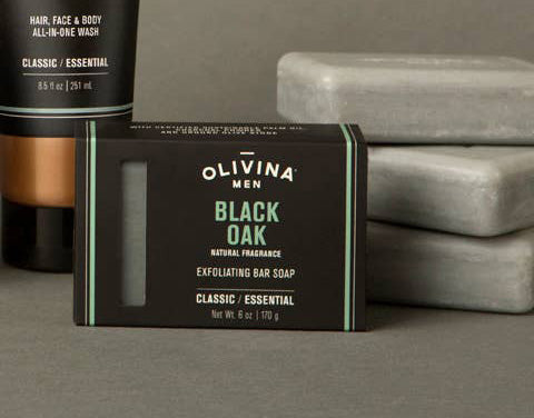 A bar of Barrel and Oak Black Oak Exfoliating Bar Soap is showcased in its sleek black and green packaging. Behind it, gray bars of soap and a bottle of all-in-one wash are visible, each crafted with sustainable palm oil to ensure eco-friendly grooming.