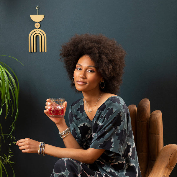 A woman with natural curly hair sits in a wooden chair, holding a drink. She wears a patterned dress and hoop earrings. Behind her, the modern space features a Scout Jewelry Nebula Rose Quartz Wall Hanging and a lush green plant.
