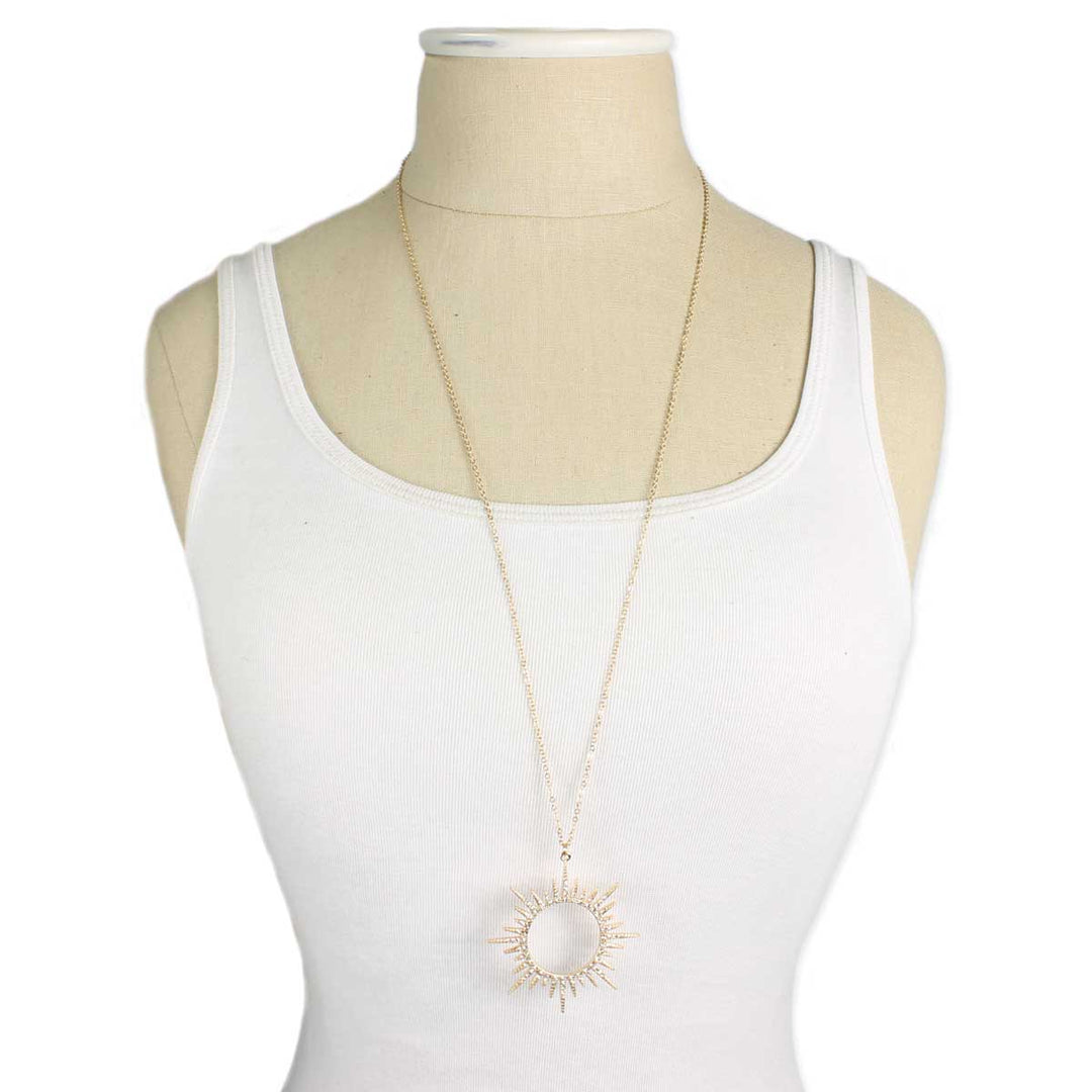 A mannequin dressed in a white tank top wears a long gold necklace from ZAD Jewelry, featuring the Gold Sunburst Crystal Necklace with a gold plate finish. The pendant, positioned at chest level, has a circular center with rays extending outward on a fine gold link chain. The tank top and necklace create a simple yet elegant display.