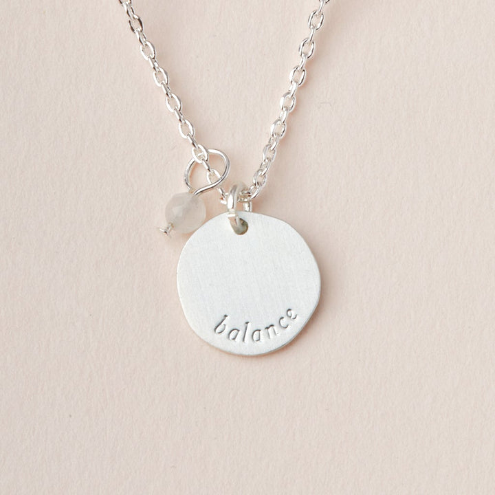 The Intention Neck Moonstone/Silver by Scout Jewelry showcases a round pendant inscribed with the word "balance" and adorned with semi-precious gemstones. It is elegantly displayed on an adjustable brass chain, highlighted by a small, translucent bead set against a soft beige background.