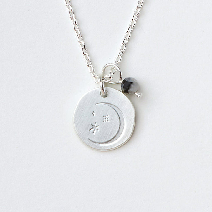 The Intention Neck Moonstone/Silver by Scout Jewelry is a silver necklace adorned with etched icon charms, including a circular pendant featuring an engraved crescent moon and star. Complementing it, a small dark bead and a silver heart charm gracefully hang from an adjustable brass chain against a simple, light-colored background.