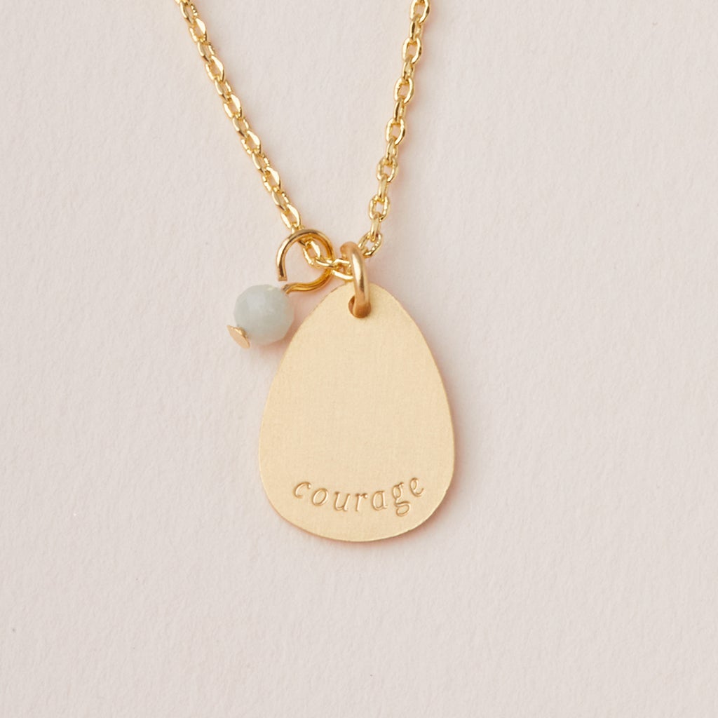 The Intention Neck Amazonite/Gold by Scout Jewelry showcases a teardrop-shaped pendant engraved with "courage," accented by a small light blue bead like semi-precious gemstones, elegantly hanging from an adjustable chain against a light-colored background.