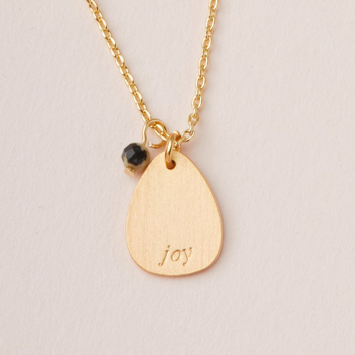 The Scout Jewelry Intention Neck Dalmatian/Gold features a teardrop pendant engraved with "joy" and a black bead charm, both on an adjustable gold chain.