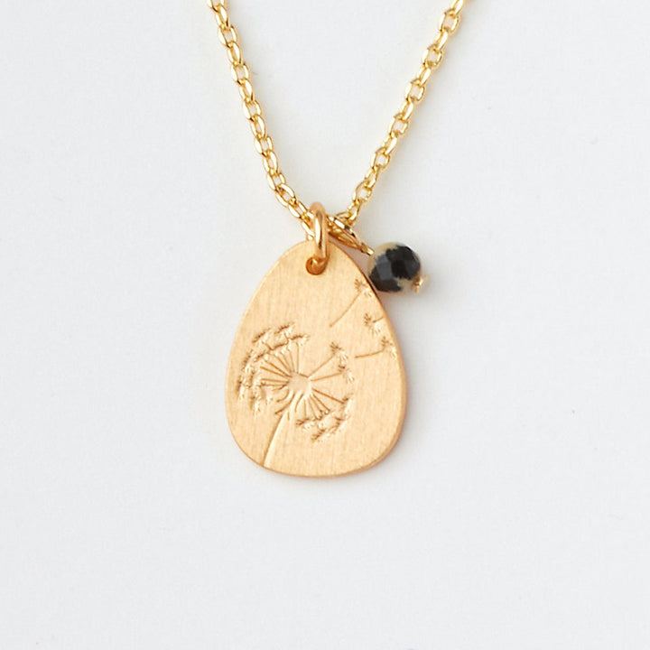 The Intention Neck Dalmatian/Gold by Scout Jewelry features a gold pendant necklace with a dandelion engraving on an adjustable chain, complemented by a small black bead charm. Set against a plain white background.