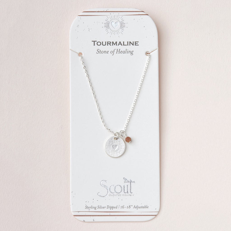 The "Intention Neck Tourmaline/Silver" by Scout Jewelry is a silver necklace with a round pendant and small stone, packaged on a "Tourmaline Stone of Healing" card. It features semi-precious gemstones, an engraved charm, and an adjustable 16-18 inch sterling silver dipped chain.