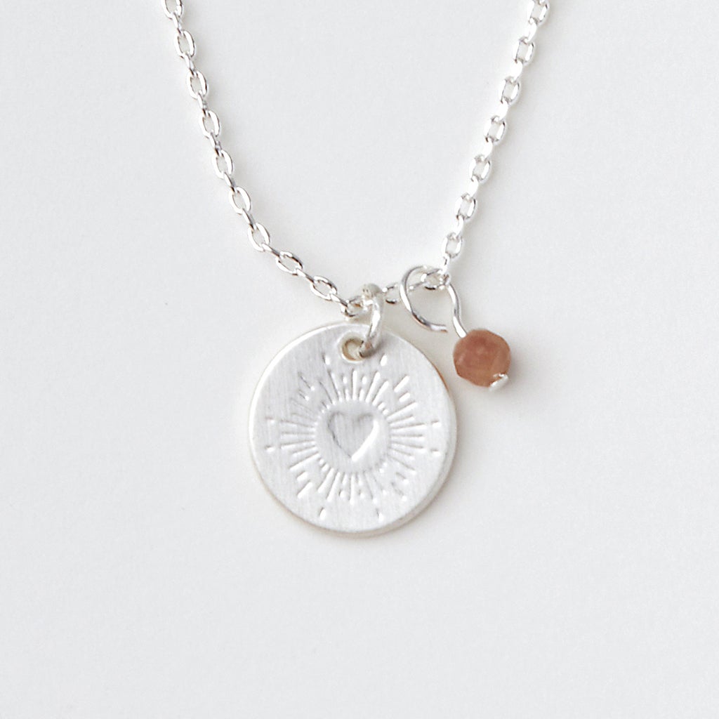 The Scout Jewelry Intention Neck Tourmaline/Silver is a delicate silver necklace with a round pendant engraved with heart and sunburst charms. A small brown bead hangs beside it, both on an adjustable chain against a plain background.