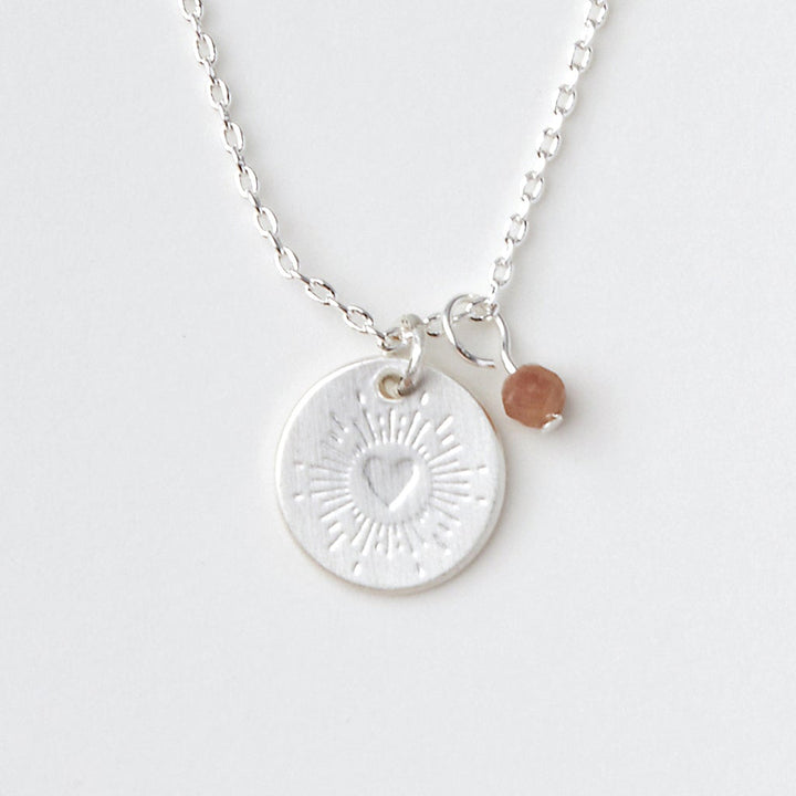 The Scout Jewelry Intention Neck Tourmaline/Silver is a delicate silver necklace with a round pendant engraved with heart and sunburst charms. A small brown bead hangs beside it, both on an adjustable chain against a plain background.