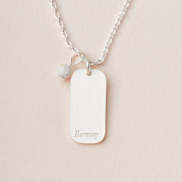 The Intention Neck Howlite/Silver by Scout Jewelry is a beautiful silver necklace featuring an adjustable chain with a rectangular pendant and a small bead charm. The engraved charm prominently displays the word "Harmony." This delicate piece is elegantly showcased against a plain off-white background.