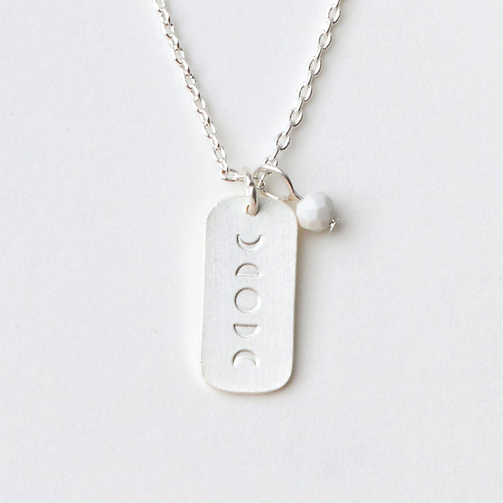 A close-up image of the Scout Jewelry "Intention Neck Howlite/Silver" necklace featuring a rectangular pendant with cut-out moon phases and a small white bead attached to the adjustable chain. The necklace is displayed on a plain, light-colored background, highlighting its simplicity and elegance.