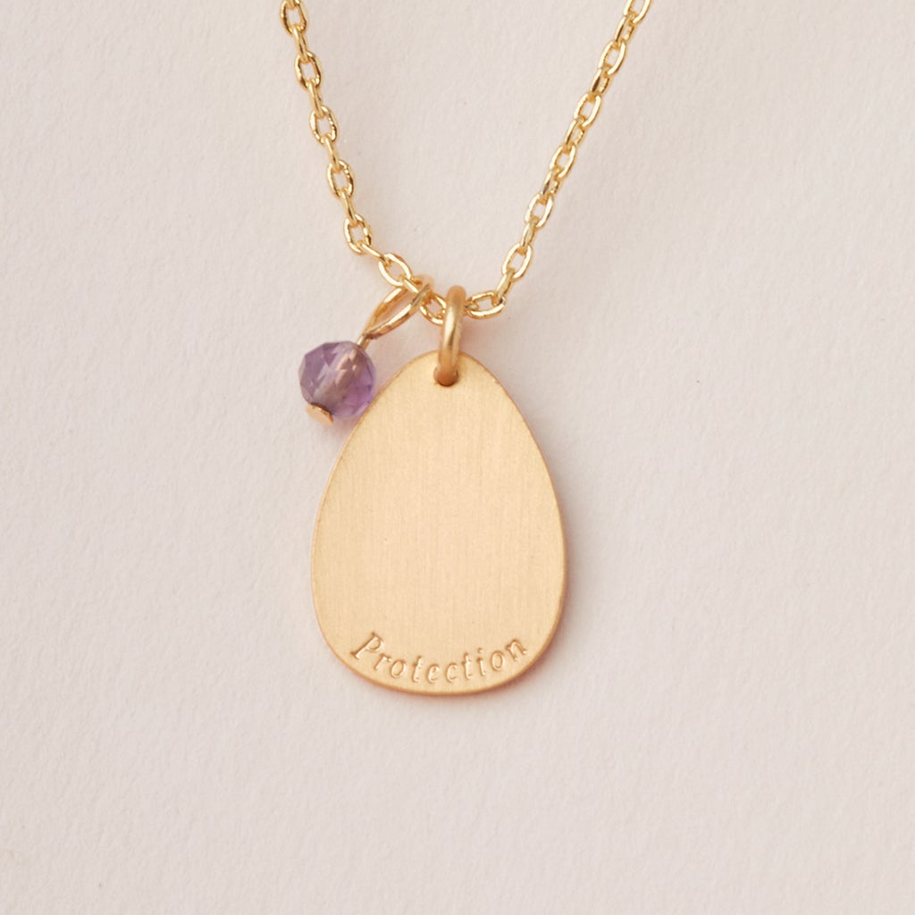 The Intention Neck Amethyst/Gold necklace by Scout Jewelry features a teardrop-shaped engraved charm with the word "Protection" on an adjustable brass chain. Attached to the pendant is a small purple semi-precious stone, and it is displayed against a neutral background.