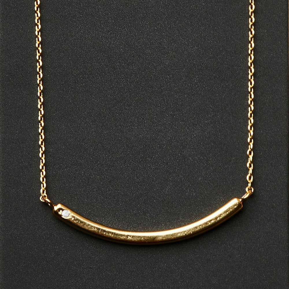 The Refined Neck Comet/Gold by Scout Jewelry is a stunning piece, featuring a curved bar pendant adorned with two small diamonds on one side. This exquisite pendant elegantly rests against a dark background and is suspended horizontally from a delicate 18k gold vermeil chain, enhancing its luxurious appeal.