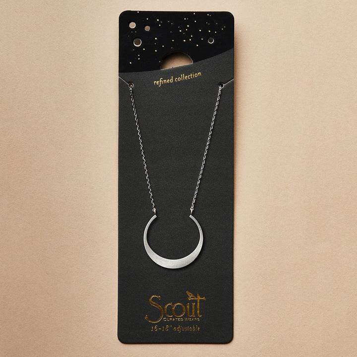 A sterling silver crescent moon necklace from Scout Jewelry's "Refined Neck Crescent/Silver" collection is elegantly displayed on a black card adorned with gold stars and the words "refined collection." The brand name "Scout Jewelry" is highlighted in gold lettering at the bottom, along with "15-18 adjustable." Set against a plain light tan background, it’s an exquisite piece from their elegant line.