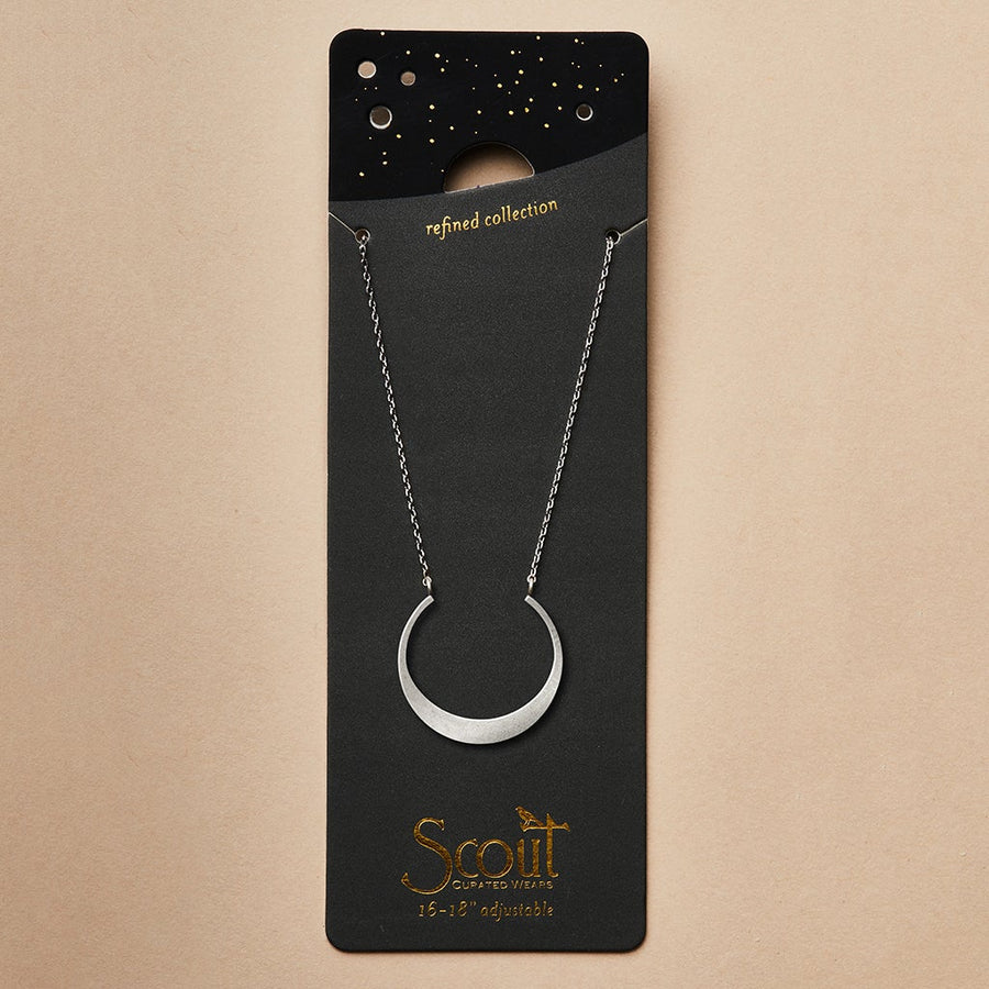 A sterling silver crescent moon necklace from Scout Jewelry's "Refined Neck Crescent/Silver" collection is elegantly displayed on a black card adorned with gold stars and the words "refined collection." The brand name "Scout Jewelry" is highlighted in gold lettering at the bottom, along with "15-18 adjustable." Set against a plain light tan background, it’s an exquisite piece from their elegant line.