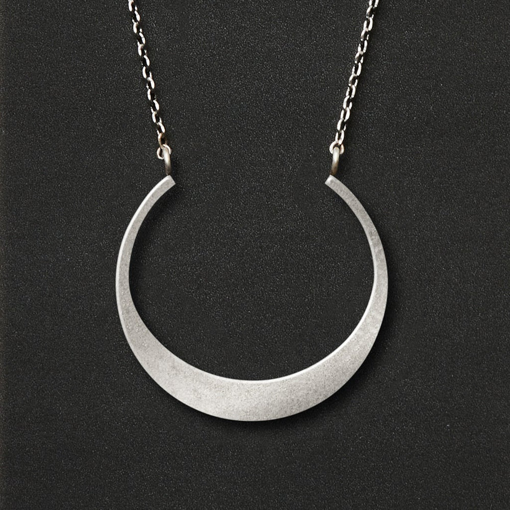 The Refined Neck Crescent/Silver by Scout Jewelry features a crescent-shaped pendant crafted from sterling silver, beautifully showcased on a delicate chain against a dark backdrop.