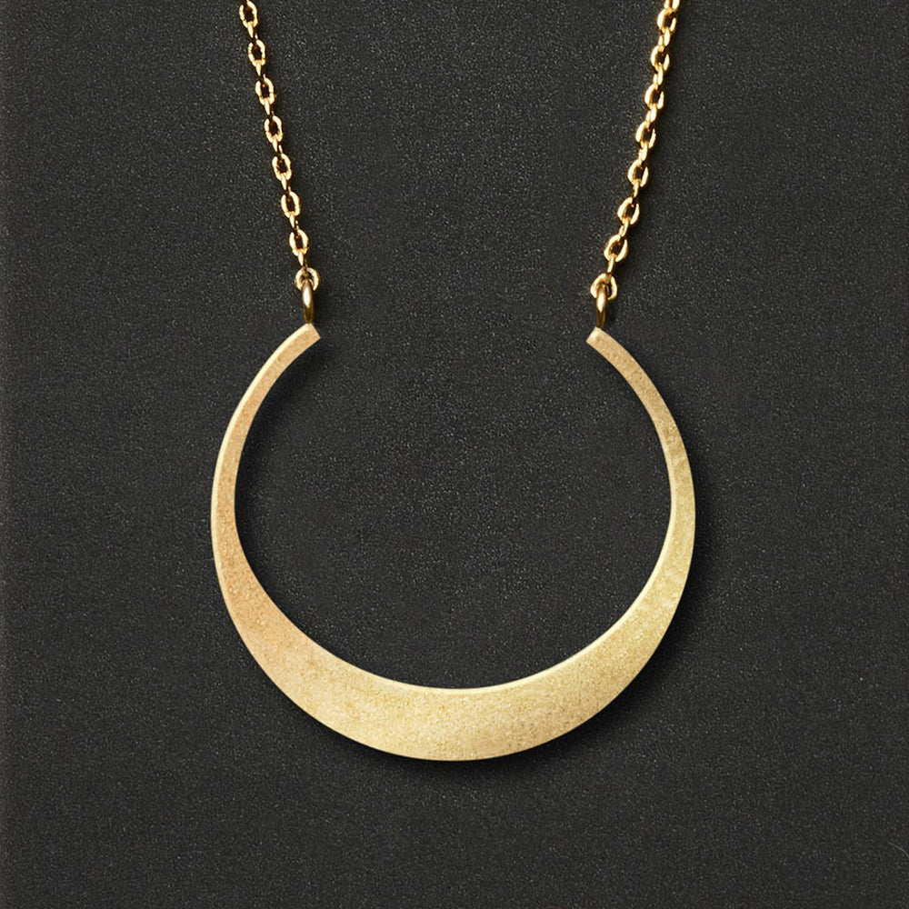 A Refined Neck Crescent/Gold necklace from Scout Jewelry, featuring a crescent-shaped pendant with a smooth and shiny 18k gold vermeil finish, hangs elegantly from an adjustable chain against a dark background.