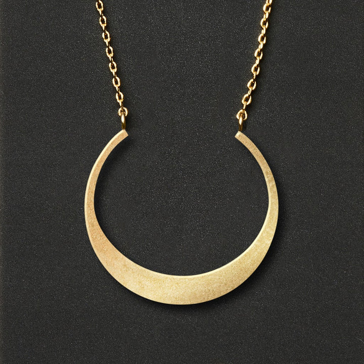 A Refined Neck Crescent/Gold necklace from Scout Jewelry, featuring a crescent-shaped pendant with a smooth and shiny 18k gold vermeil finish, hangs elegantly from an adjustable chain against a dark background.