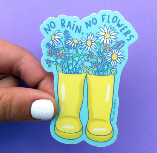 A vibrant and durable vinyl sticker from Lucky Sardine, called the "No Rain No Flowers Sticker," showcases two yellow rain boots filled with a variety of flowers against a purple backdrop. The phrase "No Rain, No Flowers" is elegantly curved above in a blue font. A hand with white nail polish proudly holds this weatherproof sticker.