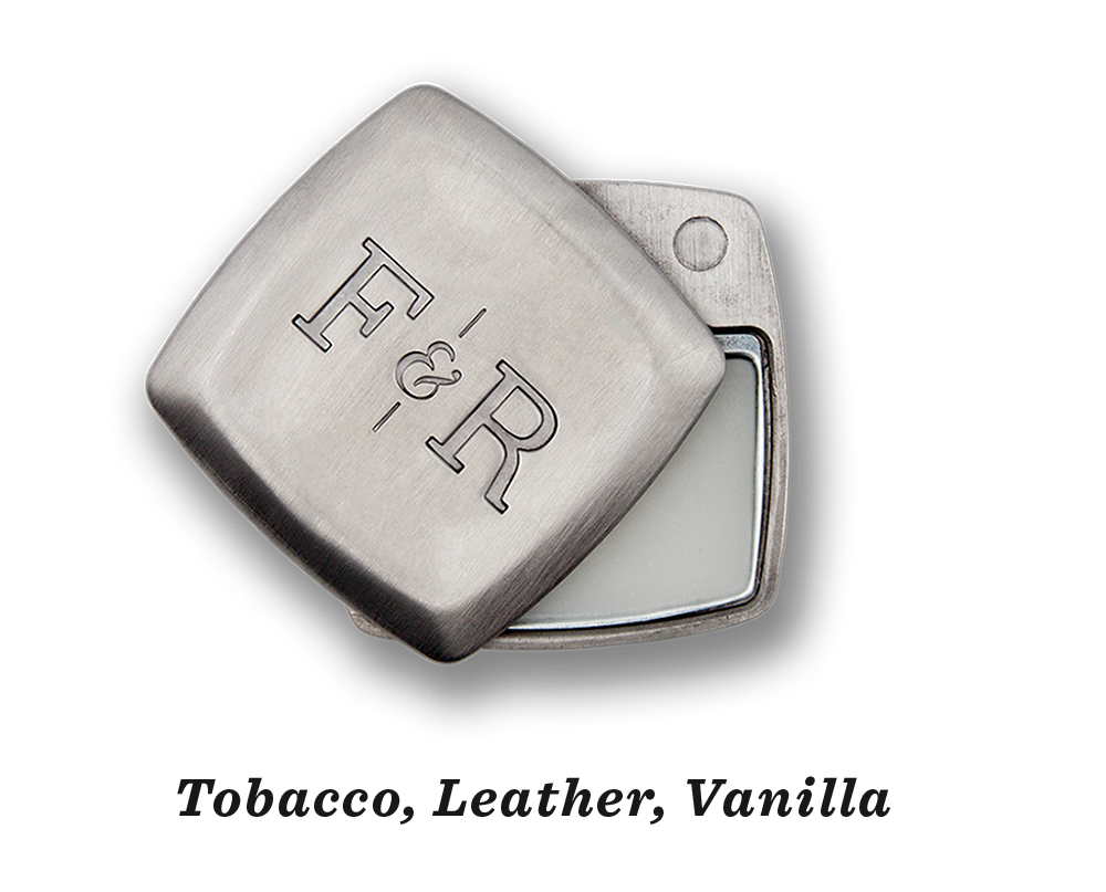 The image displays a compact, square metal tin from Fulton & Roark, embossed with "F&R" on the lid. The partially opened lid reveals a white balm inside, emitting a vanilla scent. This is their Sterling .2oz product.