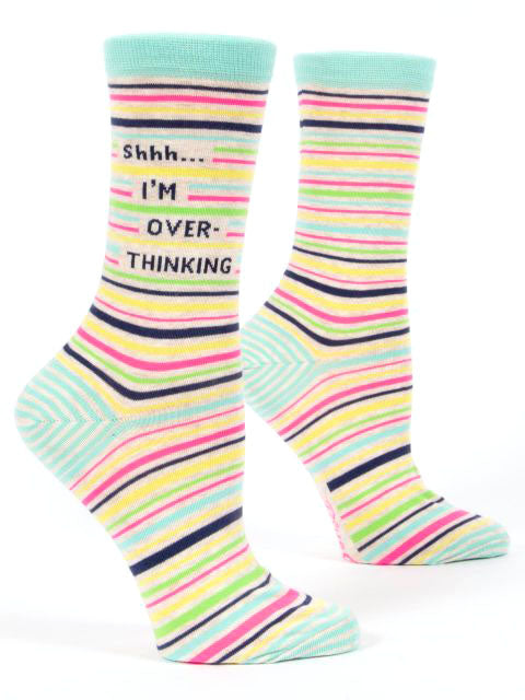 The "Shhhh Overthinking" socks from Blue Q are a pair of colorful striped socks featuring light blue, pink, yellow, and black lines. One sock displays the text "Shhh... I'M OVER-THINKING" written on the side in black letters. Made from a blend of nylon, combed cotton, and spandex, these socks fit women's shoe sizes 5-10 and have light blue cuffs, heels, and toes.